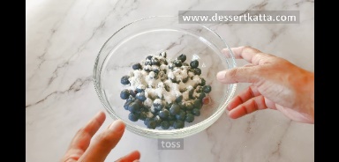 a glass bowl contains blueberries and all-purpose flour.