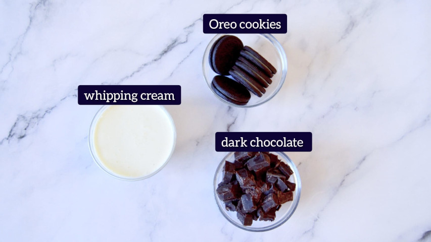 Oreo cookies, dark chocolate and whipping cream are placed in small glass bowls to make topping for mini Oreo cheesecake